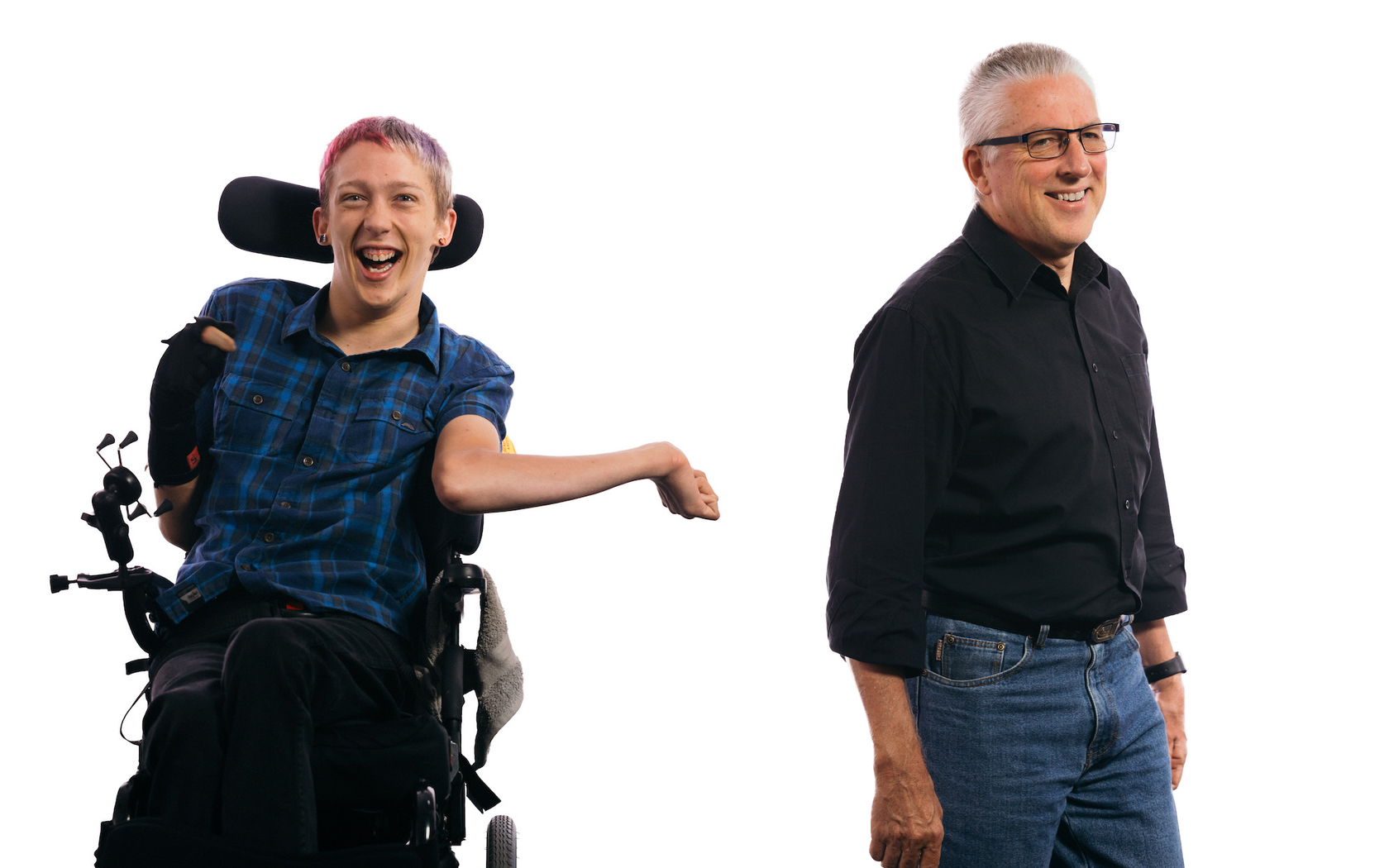 A middle-aged man wearing glasses stands and smiles off-camera. Next to him a young man with faded red and blue hair, seated in a wheelchair, smiles to camera.
